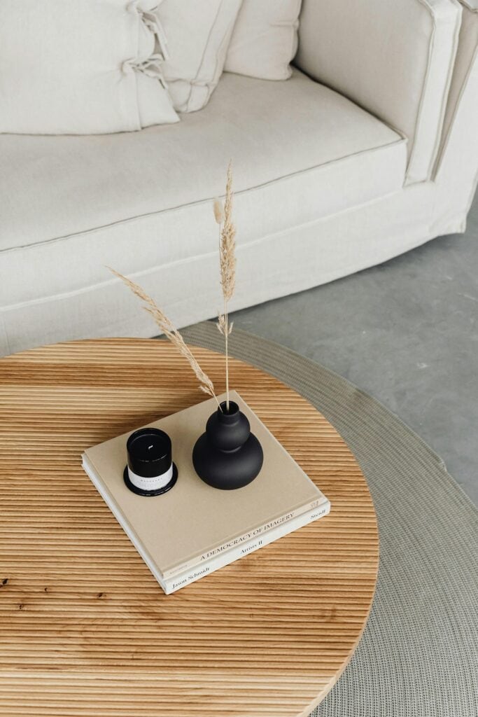 A round wooden coffee table with two books, a small black vase with dried grass, and a black candle, in front of a light-colored sofa.