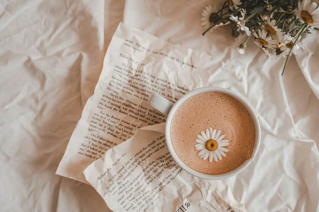 A cup of frothy coffee with a daisy flower on top sits on two torn book pages, with more daisies nearby, all placed on a crumpled white fabric surface.