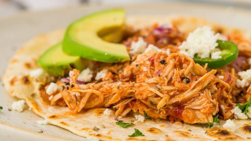 A close-up of a soft tortilla topped with shredded chicken, sliced avocado, crumbled cheese, chopped herbs, and red onions—perfect for easy dinners and those who love delicious slow cooker recipes.