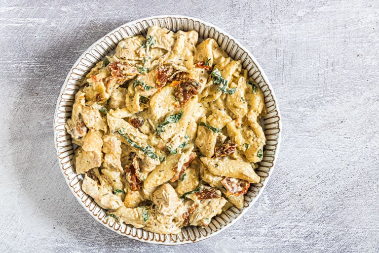 A bowl of creamy pasta with sun-dried tomatoes and spinach, served in a patterned dish on a light textured surface—one of those easy meals perfect for Crockpot dinners.
