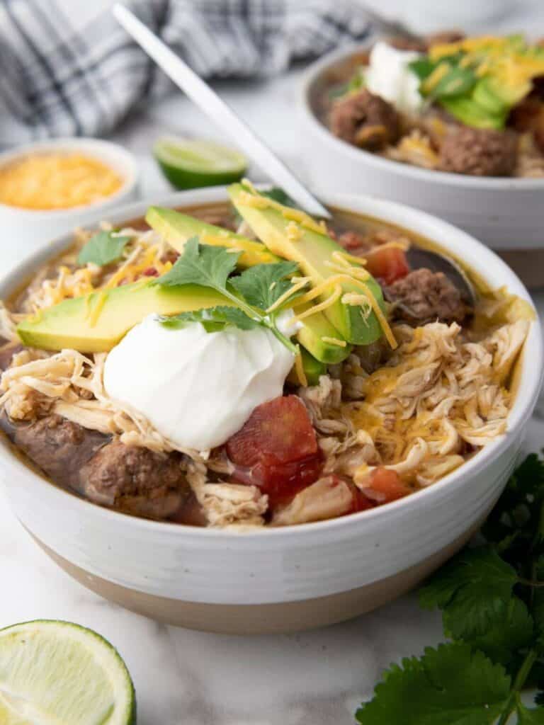 A bowl of shredded chicken and beef soup topped with avocado slices, shredded cheese, sour cream, and cilantro—an ideal choice for Crockpot dinners. A lime wedge and extra cheese complete this easy dinner idea.