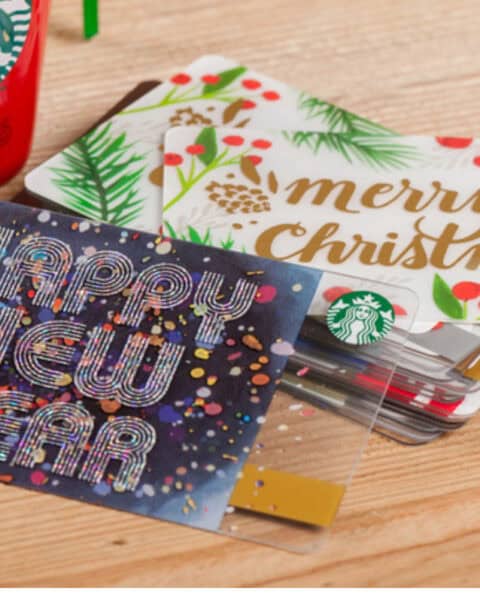 Three Starbucks gift cards with festive designs, one reading "Happy New Year" and another "Merry Christmas," are displayed on a wooden surface next to a red cupโperfect for gifting or redeeming your $5 Gift Card this season.