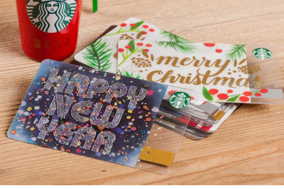 Three Starbucks gift cards with festive designs, one reading "Happy New Year" and another "Merry Christmas," are displayed on a wooden surface next to a red cup—perfect for gifting or redeeming your $5 Gift Card this season.