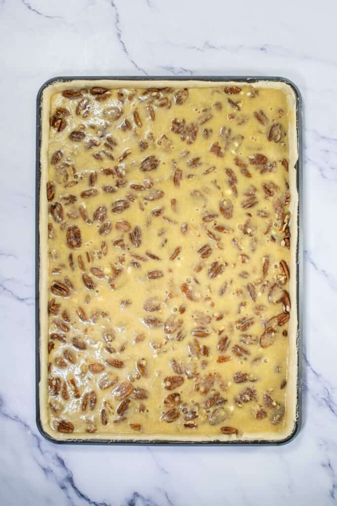 A rectangular baking pan filled with unbaked Pecan Pie Bars mixture sits on a white marble surface, ready to be transformed into an easy dessert.
