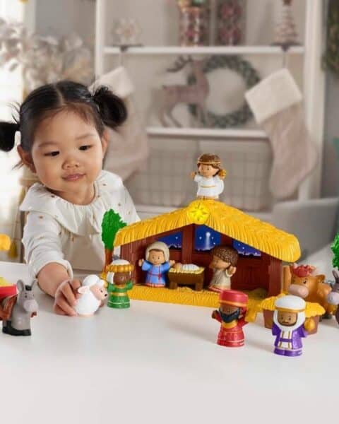 A young child plays with a Little People Nativity Set, carefully arranging the figurines of people and animals on a white table in a cozy home setting.