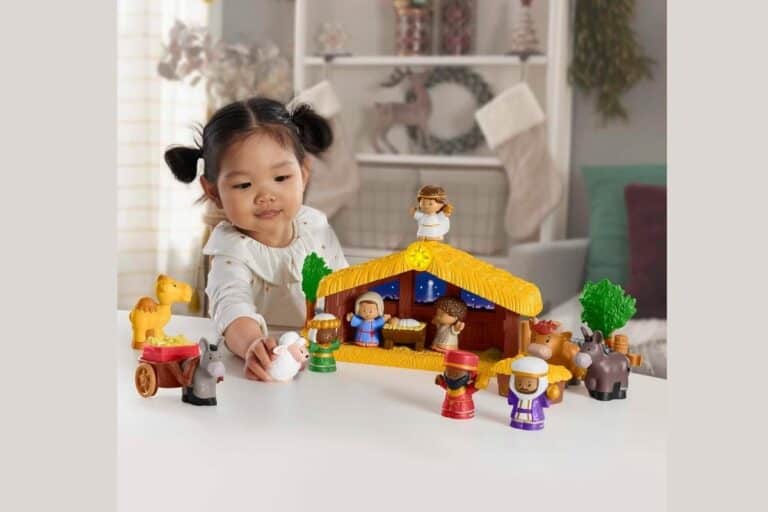 A young child plays with a Little People Nativity Set, carefully arranging the figurines of people and animals on a white table in a cozy home setting.