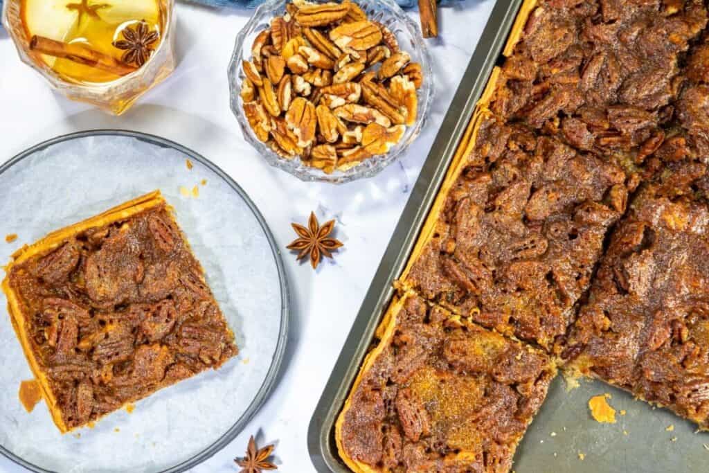 A baking tray of easy pecan pie dessert bars with one piece served on a plate, a bowl of pecans, and a glass of drink nearby on a white surface.