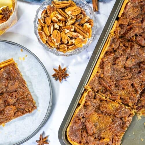 A baking tray of easy pecan pie dessert bars with one piece served on a plate, a bowl of pecans, and a glass of drink nearby on a white surface.