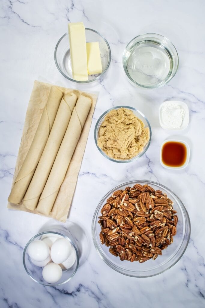 Ingredients for easy pecan pie dessert bars arranged on a marble surface, including pie crusts, eggs, pecans, butter, brown sugar, corn syrup, vanilla extract, and flour in glass bowls.
