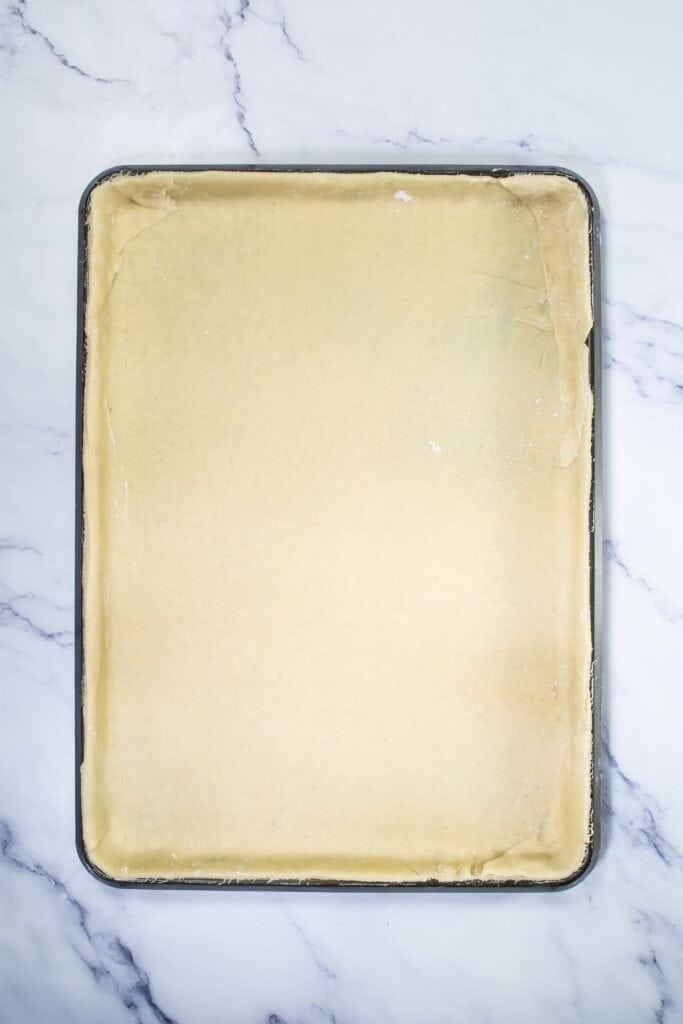 A rectangular baking sheet lined with an even layer of unbaked dough, ready to be transformed into delicious pecan pie dessert bars, sits on a white marble surface.