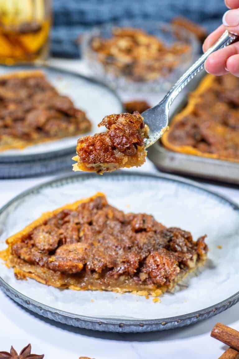 A slice of pecan pie on a plate with a fork holding a bite above it; more of this classic dessert and easy recipe pieces are visible in the background.