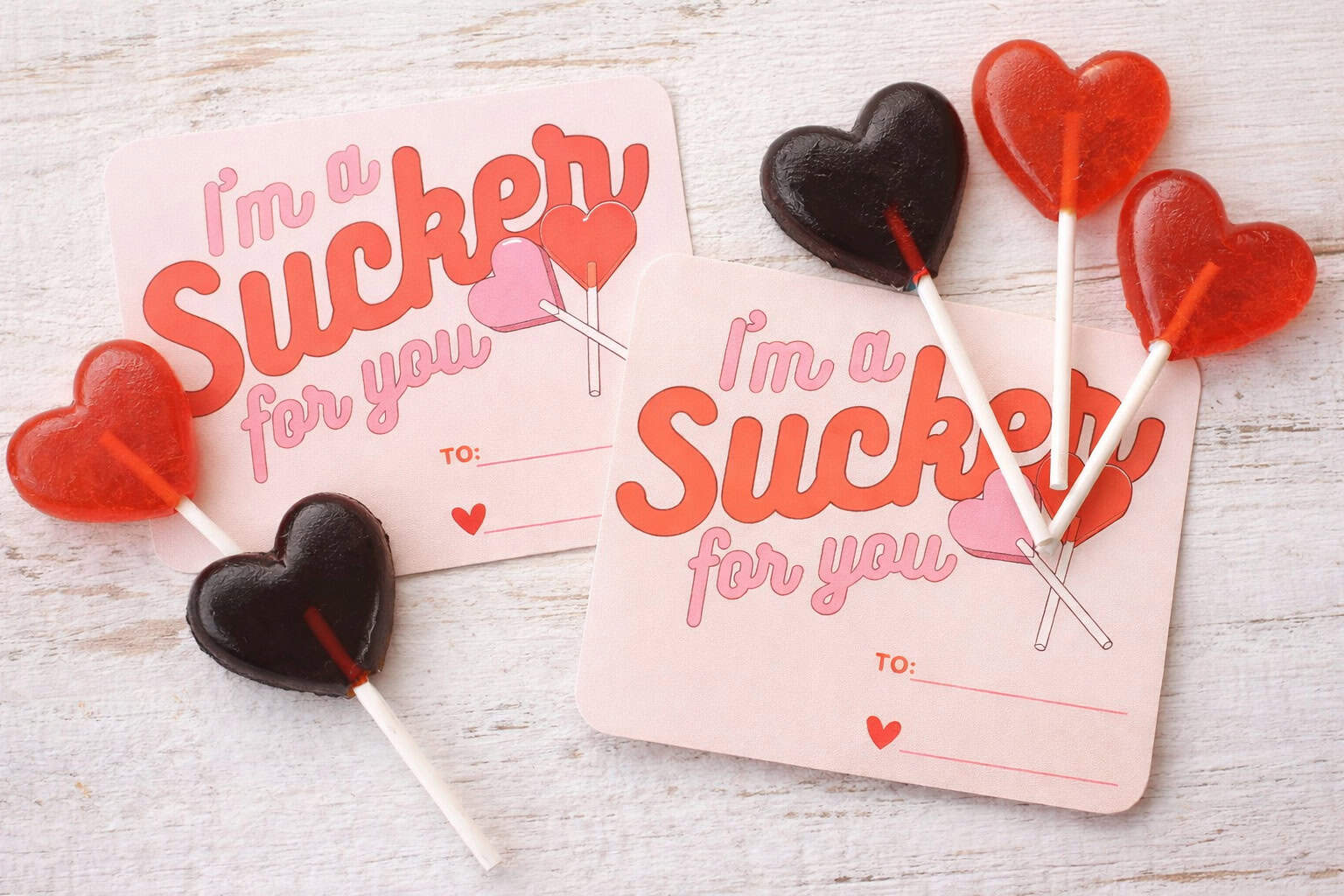 Two cards reading "I'm a Sucker for you" with blank "To" and "From" lines, each decorated with heart-shaped red and black Candy Suckers on a white wooden surface.