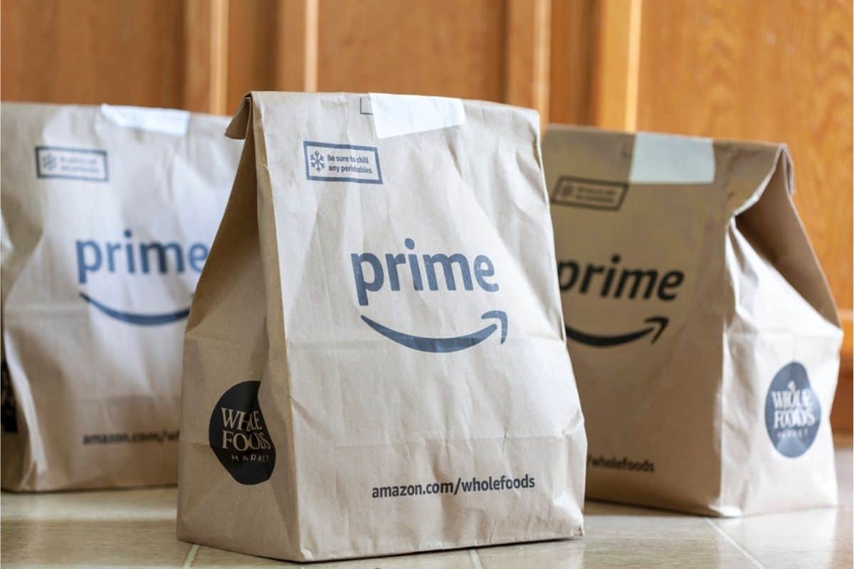 Three paper grocery bags labeled "prime" and "Whole Foods Market" sit on a tiled floor in front of wooden cabinets, showcasing fresh groceries you can get when you use EBT Amazon for easy, convenient shopping.