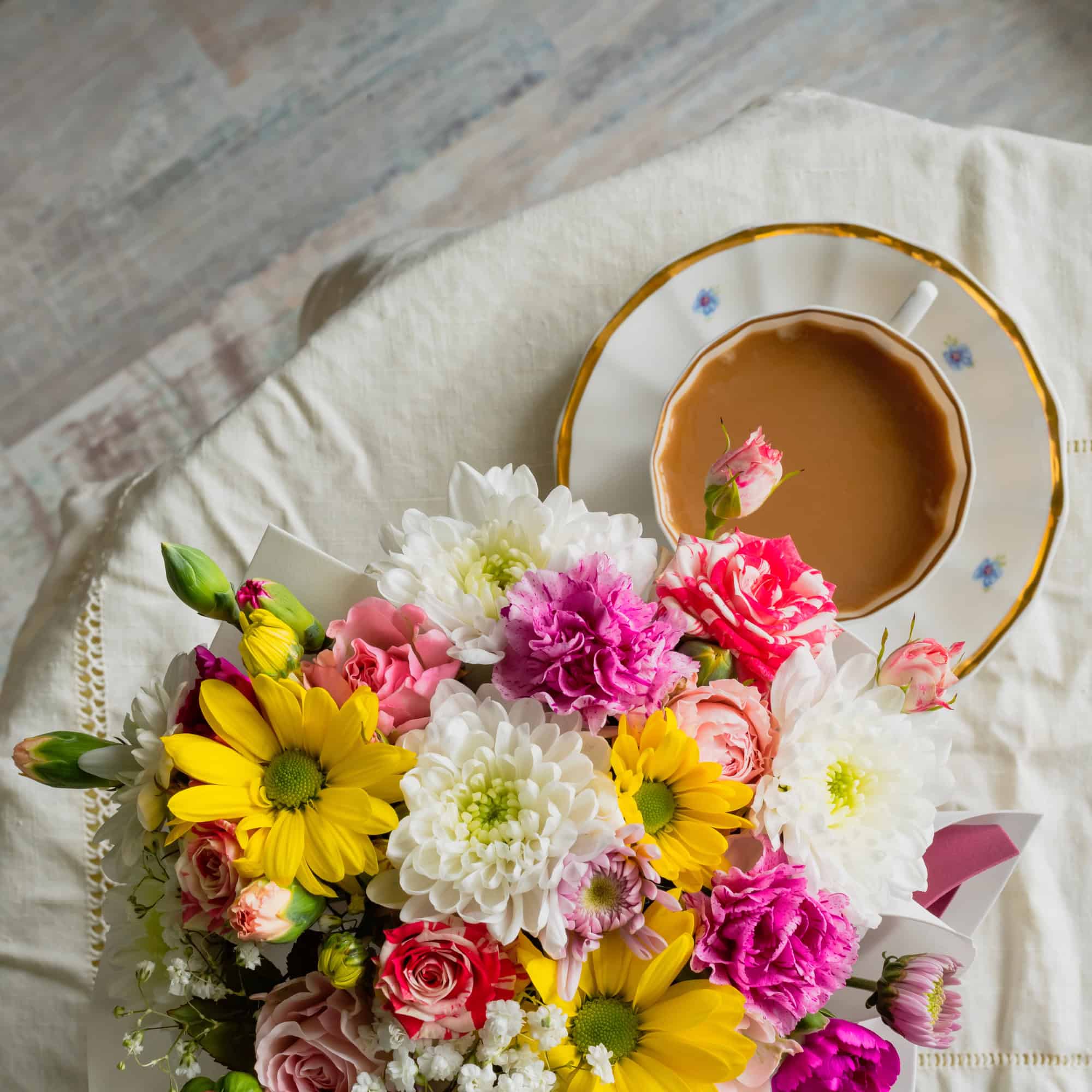 A bouquet of colorful flowers sits on a table next to a cup of coffee in a floral-patterned saucer.