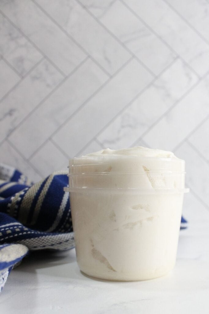 A clear plastic container filled with white cream, an easy grout cleaner, sits on a white surface next to a blue and white striped cloth with a tiled background.