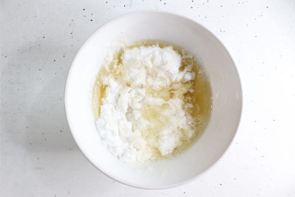 A white bowl containing a mixture of oil, liquid, and white powdery substances sits on a white speckled surface, ready to be used as an easy DIY grout cleaner.