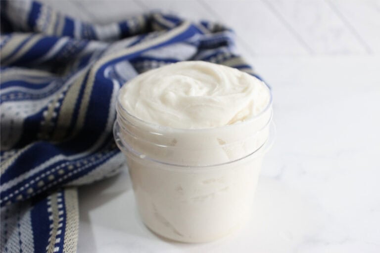 A clear plastic jar filled with a creamy, white DIY Grout Cleaner sits on a white surface next to a blue striped cloth.