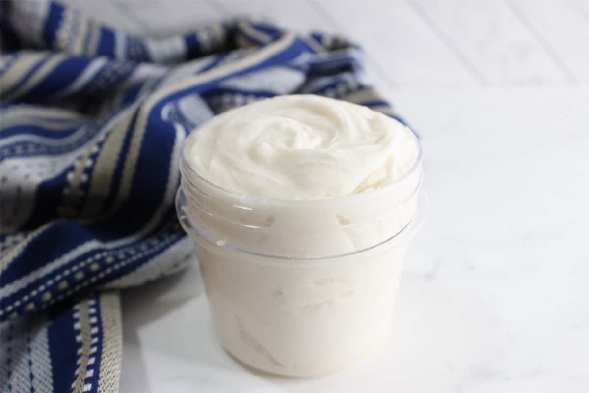 A clear plastic jar filled with a creamy, white DIY Grout Cleaner sits on a white surface next to a blue striped cloth.