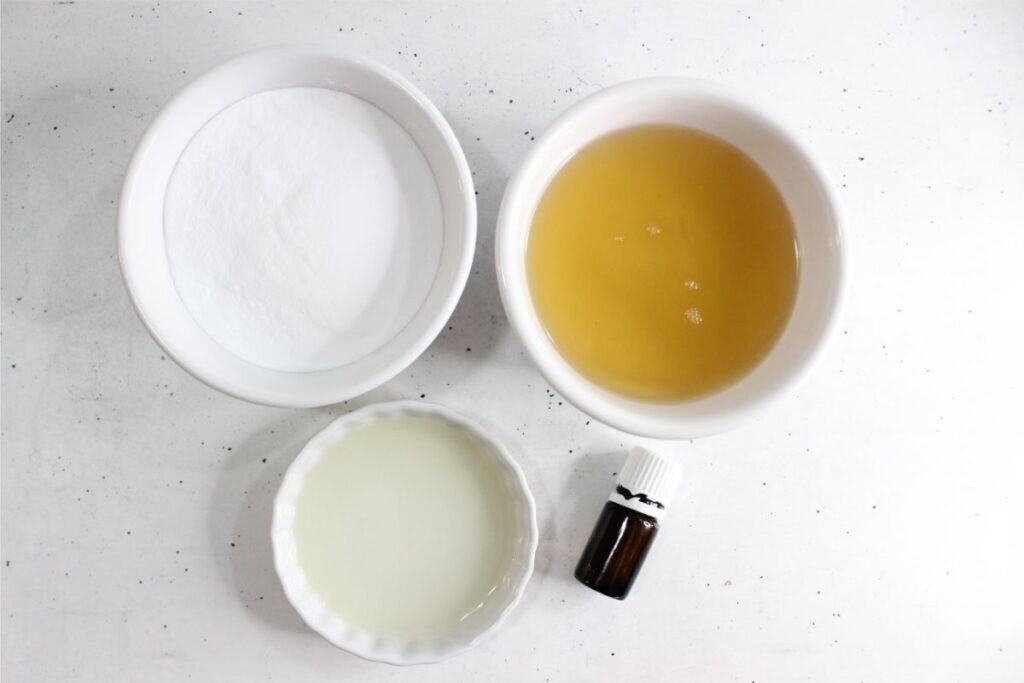 Three small bowls containing baking soda, apple cider vinegar, and lemon juice are arranged on a white surface alongside a small amber essential oil bottle—perfect ingredients for an easy DIY grout cleaner.