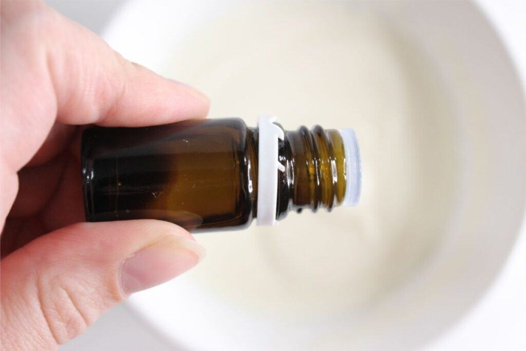 A hand holds a small amber glass bottle with an open white cap over a white bowl containing a creamy DIY Grout Cleaner.