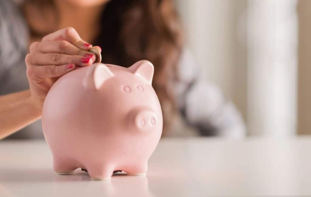 A person places a coin into a pink piggy bank on a white surface, ready to start saving and stay committed to their savings challenge.