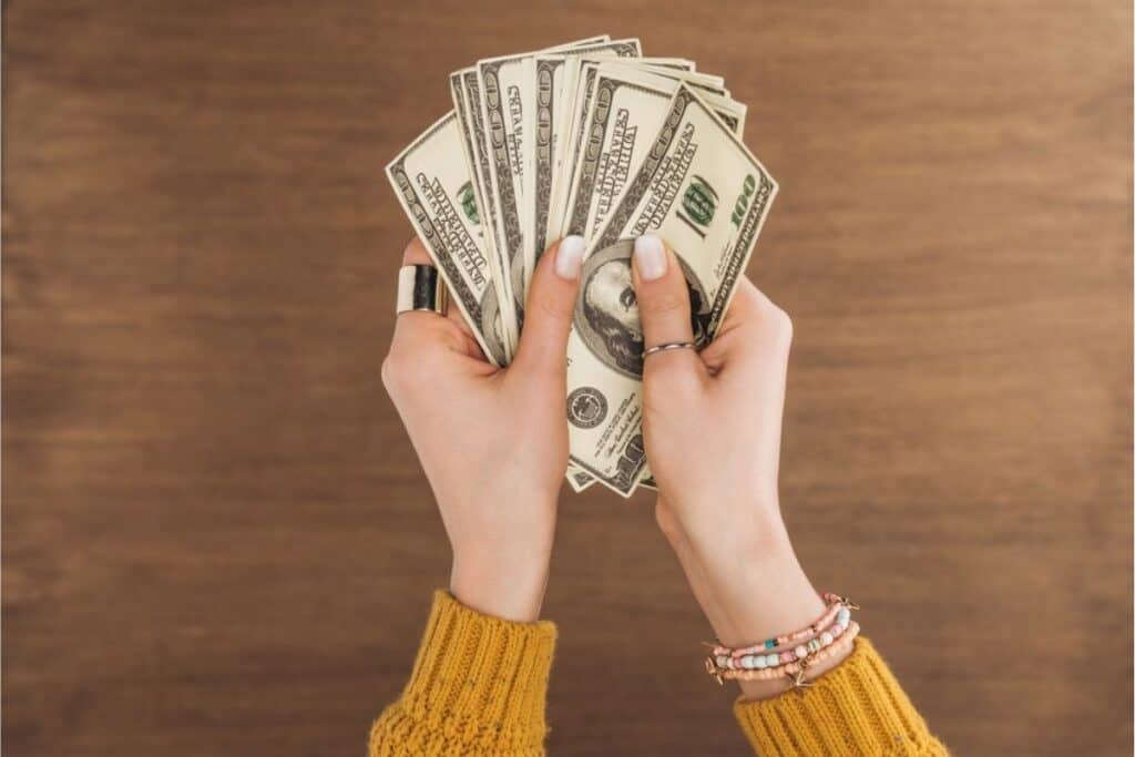 A person wearing a yellow sweater and bracelets holds several spread-out 100 US dollar bills over a wooden surface, ready to Start Saving and take on a new Savings Challenge.