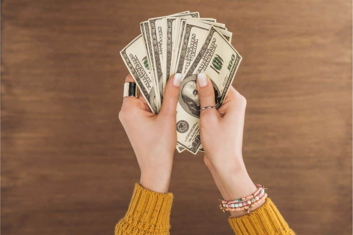 A person wearing a yellow sweater and bracelets holds several spread-out 100 US dollar bills over a wooden surface, ready to Start Saving and take on a new Savings Challenge.