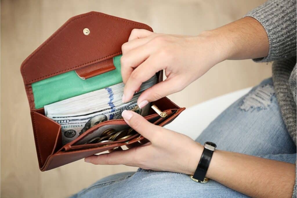 A person holds an open brown wallet overflowing with US dollar bills and coins, wearing a watch and blue jeans—ready to start a savings challenge and stay committed to building financial security.