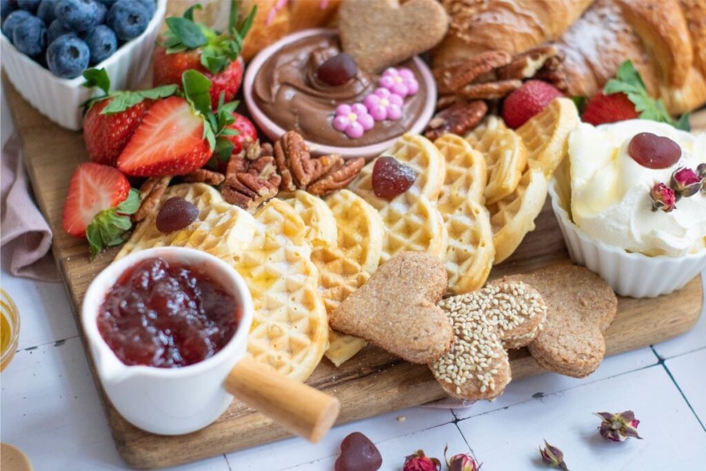 A wooden Breakfast Board with waffles, heart-shaped cookies, strawberries, blueberries, pecans, croissants, whipped cream, chocolate spread, and a small pitcher of strawberry jam—perfect for a delightful Mother's Day breakfast.