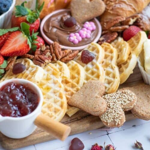 A wooden Breakfast Board with waffles, heart-shaped cookies, strawberries, blueberries, pecans, croissants, whipped cream, chocolate spread, and a small pitcher of strawberry jam—perfect for a delightful Mother's Day breakfast.