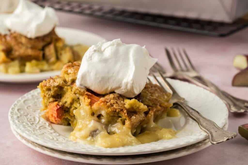 A slice of fruit pie with a golden crust, topped with whipped cream, served on a white plate with a fork; perfect for fans of seasonal desserts and the best rhubarb recipes. Another plate is in the background.