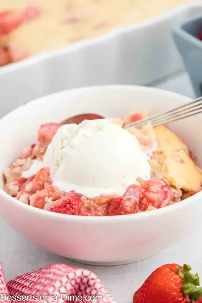 A white bowl contains strawberry cobbler topped with a scoop of vanilla ice cream and a spoon. Fresh strawberries are visible nearby, making this dessert perfect for fans of classic grandma’s rhubarb recipes.