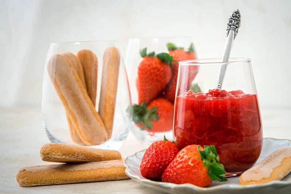 A glass of strawberry compote with a spoon, fresh strawberries, and ladyfinger biscuits arranged on a plate and in glasses—perfect for fans of seasonal desserts or anyone inspired by Grandma’s rhubarb recipes.