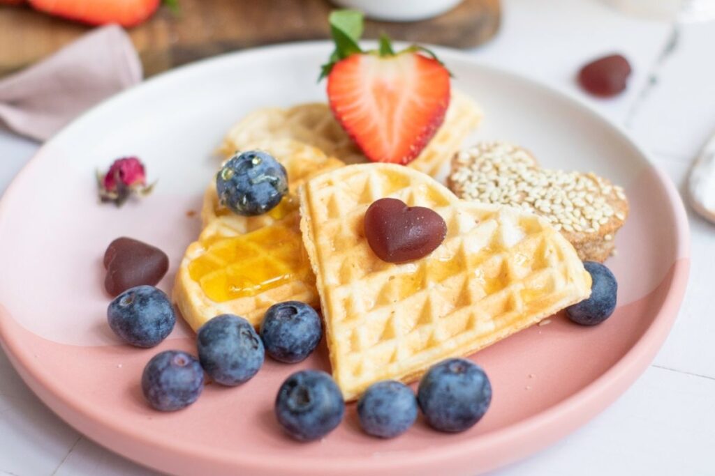 A plate with heart-shaped waffles, blueberries, a halved strawberry, jam, sesame cookie, and heart-shaped chocolate candies—perfect for a charming Mother’s Day Breakfast or as part of a delightful Breakfast Board.