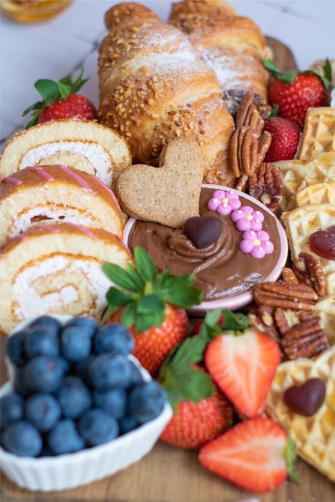 A Mother's Day Breakfast board featuring croissants, Swiss roll slices, strawberries, blueberries, waffles, pecans, and a bowl of chocolate dip topped with sprinkles and a heart-shaped cookie—perfect for celebrating Mom.