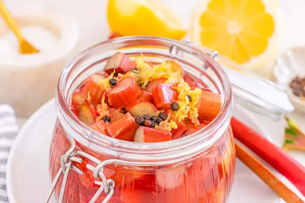 A glass jar filled with chopped rhubarb, lemon zest, and spices hints at delicious rhubarb recipes in the making, with sliced lemon and cinnamon sticks in the background.