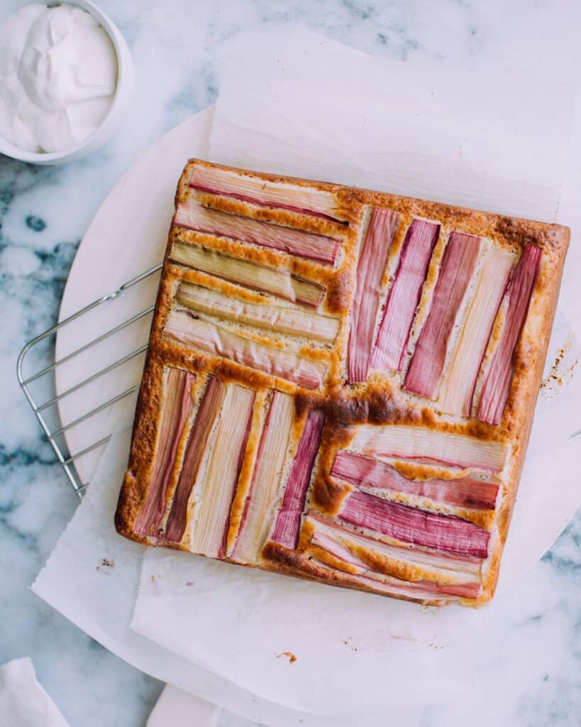 A square rhubarb cake with striped rhubarb stalks on top, resting on parchment paper and a cooling rack, with a bowl of cream nearby—perfect for fans of seasonal desserts or those seeking inspiration from grandma's recipes.