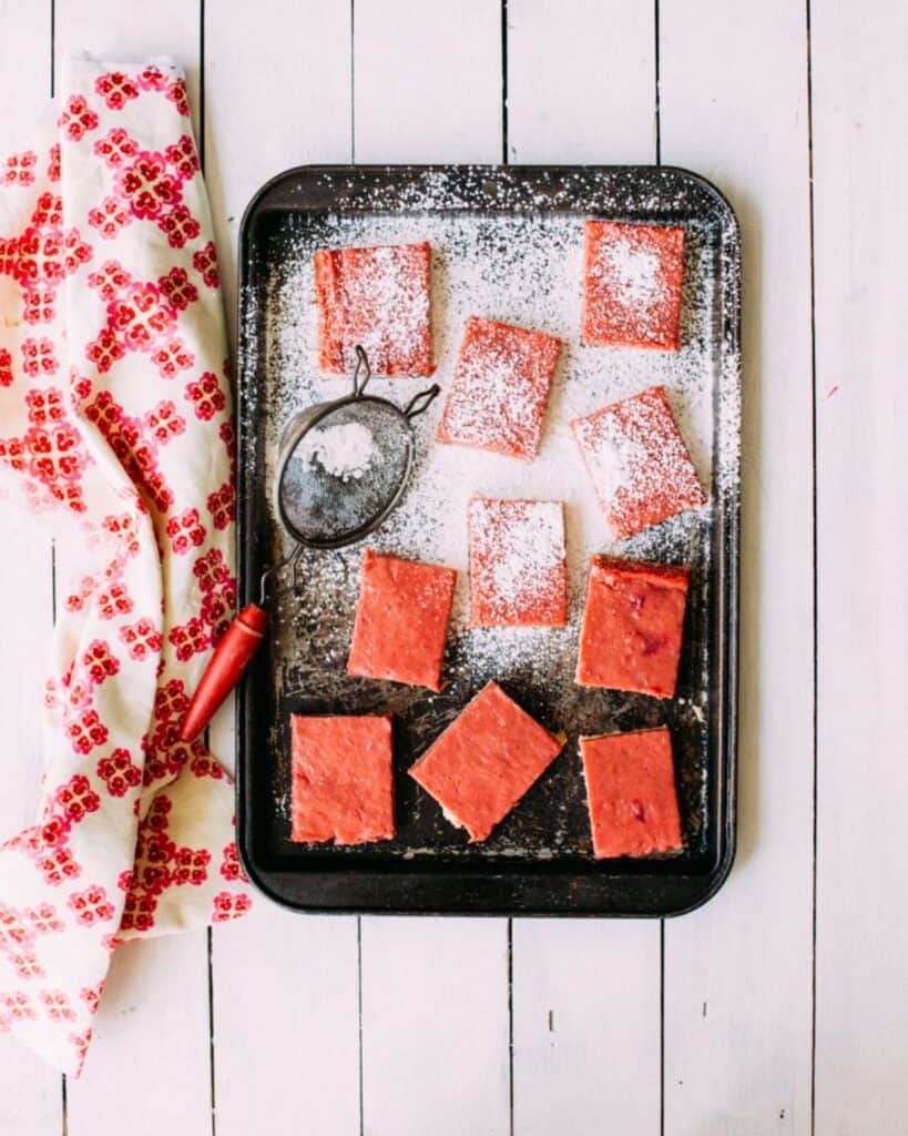 A black baking tray with pink frosted square bars, some dusted with powdered sugar, sits on a white wooden surface next to a red and white patterned cloth—perfect for those who love homemade rhubarb recipes.