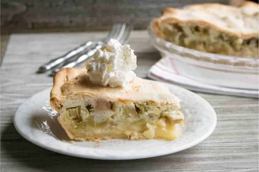 A slice of pie with a golden crust and a fruit filling is topped with whipped cream, served on a white plate with a fork. The rest of the pie is in the background.
