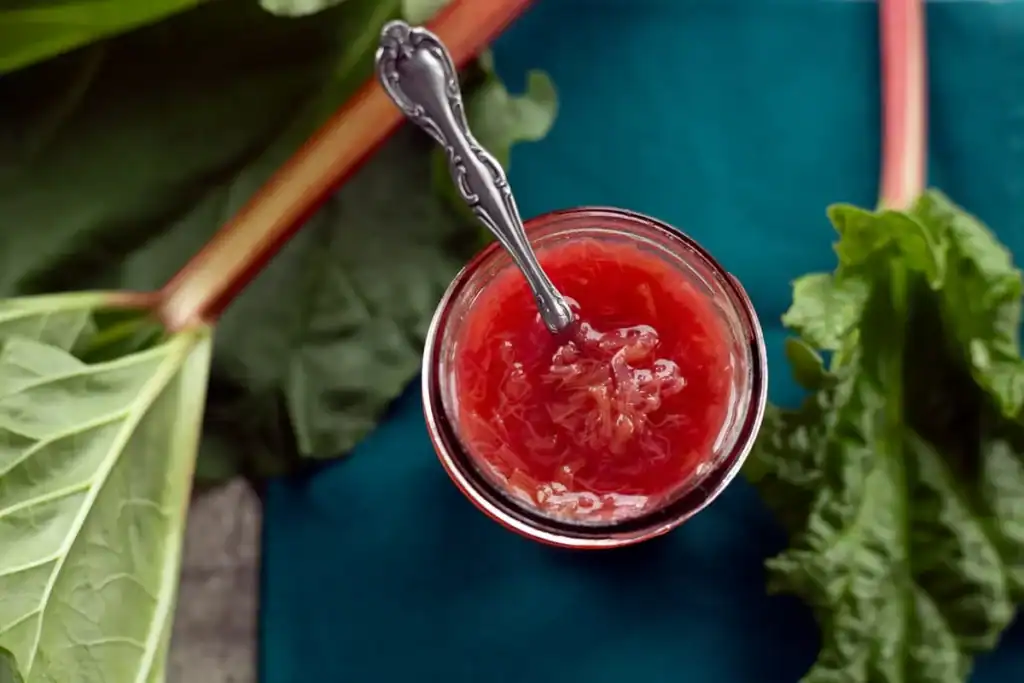 A jar of rhubarb jam with a spoon inside, surrounded by fresh rhubarb stalks and leaves on a teal surface—perfect inspiration for seasonal desserts or adding a touch of nostalgia from grandma’s recipes.
