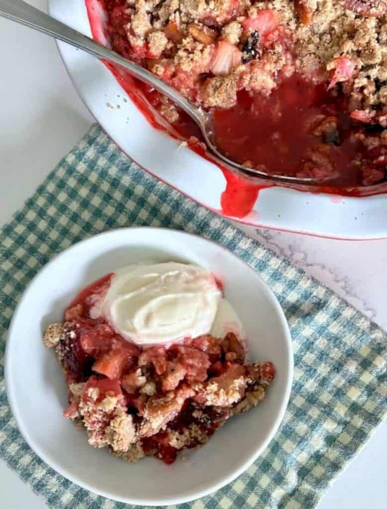 A bowl of fruit crumble—one of grandma’s classic recipes—topped with vanilla ice cream sits next to a partially served baking dish on a blue and white checkered cloth.