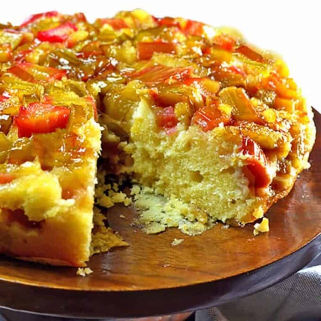 A round upside-down cake with a topping of chopped rhubarb, missing one slice, displayed on a wooden cake stand—one of the timeless rhubarb recipes perfect for seasonal desserts.