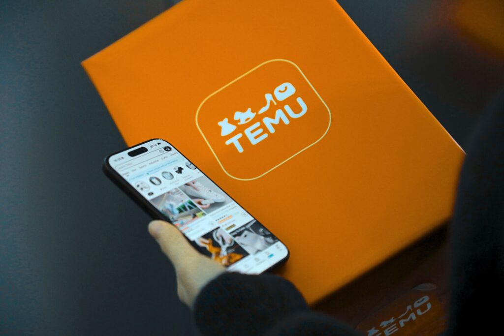 A person holds a smartphone showing the Temu shopping app beside an orange Temu-branded box, highlighting the convenience of shopping with Temu.