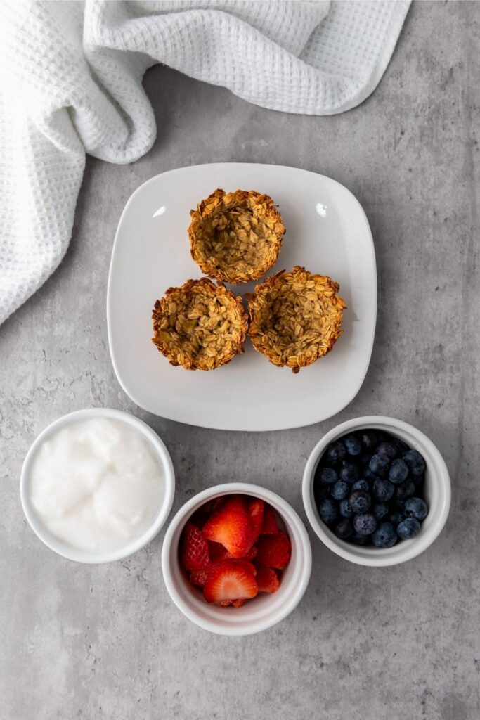Three Berry Granola Cups on a white plate, with small bowls of yogurt, sliced strawberries, and blueberries on a gray surface next to a white textured cloth. Perfect for serving with mixed berry toppings.
