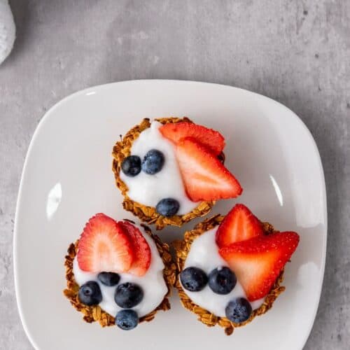 Three granola cups topped with yogurt, sliced strawberries, and blueberries are arranged on a white square plate—a delightful Berry Snacks selection for any time of day.