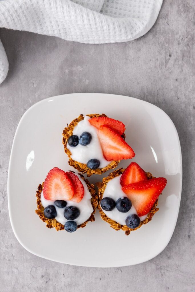 Three granola cups topped with yogurt, sliced strawberries, and blueberries are arranged on a white square plate—a delightful Berry Snacks selection for any time of day.