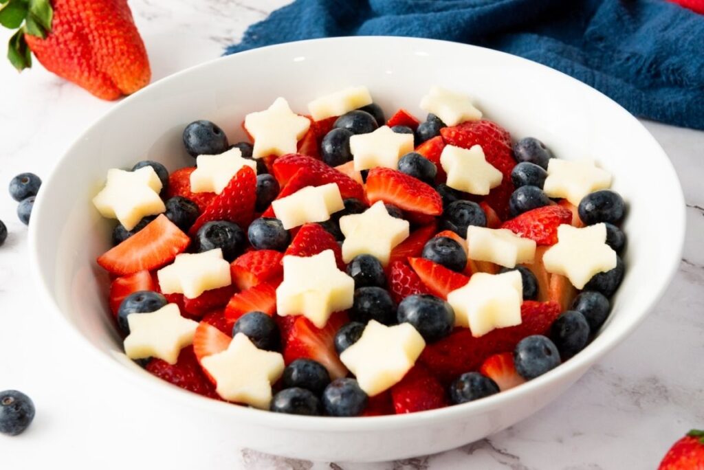 A white bowl filled with blueberries, sliced strawberries, and star-shaped pieces of white fruit creates a vibrant Red White Blue Fruit Salad on a marble surface.