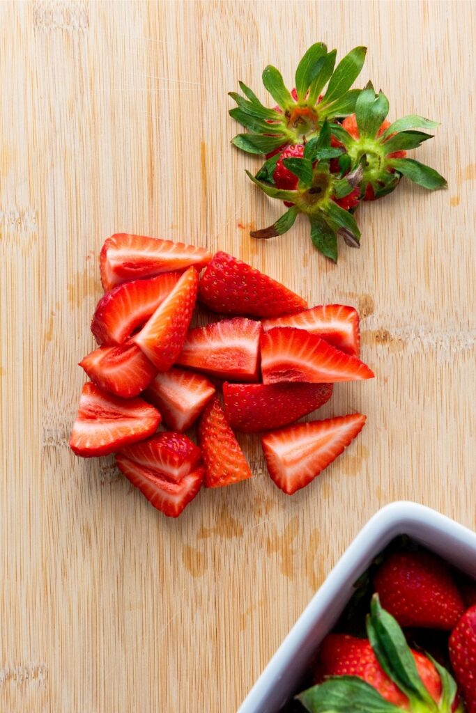 Sliced strawberries and a whole strawberry with leaves on a wooden cutting board, with part of a white container holding more strawberries in the lower right corner—perfect for adding to your favorite Red White and Blue Fruit Salad.