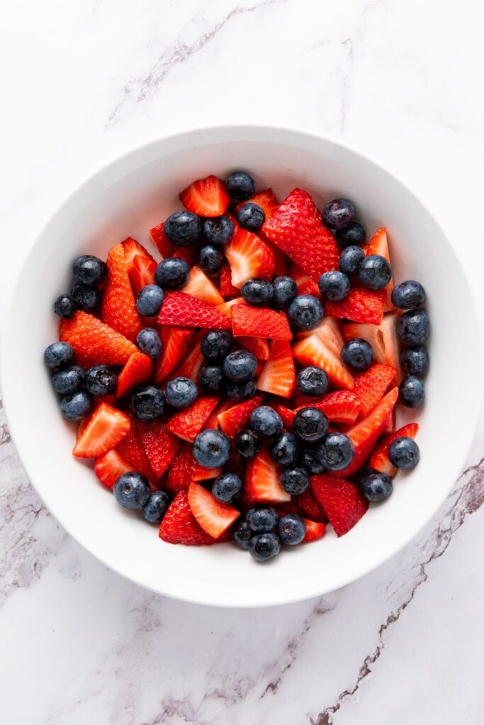 A white bowl filled with sliced strawberries and whole blueberries sits on a white marble surface, creating a vibrant Red White and Blue Fruit Salad.