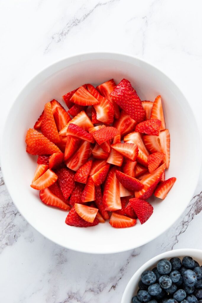 A white bowl filled with sliced strawberries sits on a marble surface, with a smaller bowl of blueberries nearby—a perfect setup for a Red White and Blue Fruit Salad.